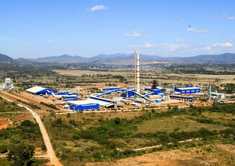 На изображении представлен промышленный объект или завод, расположенный в сельской местности или на окраине города. На заднем плане видны горы, а на переднем плане — растительность и грунтовая дорога.